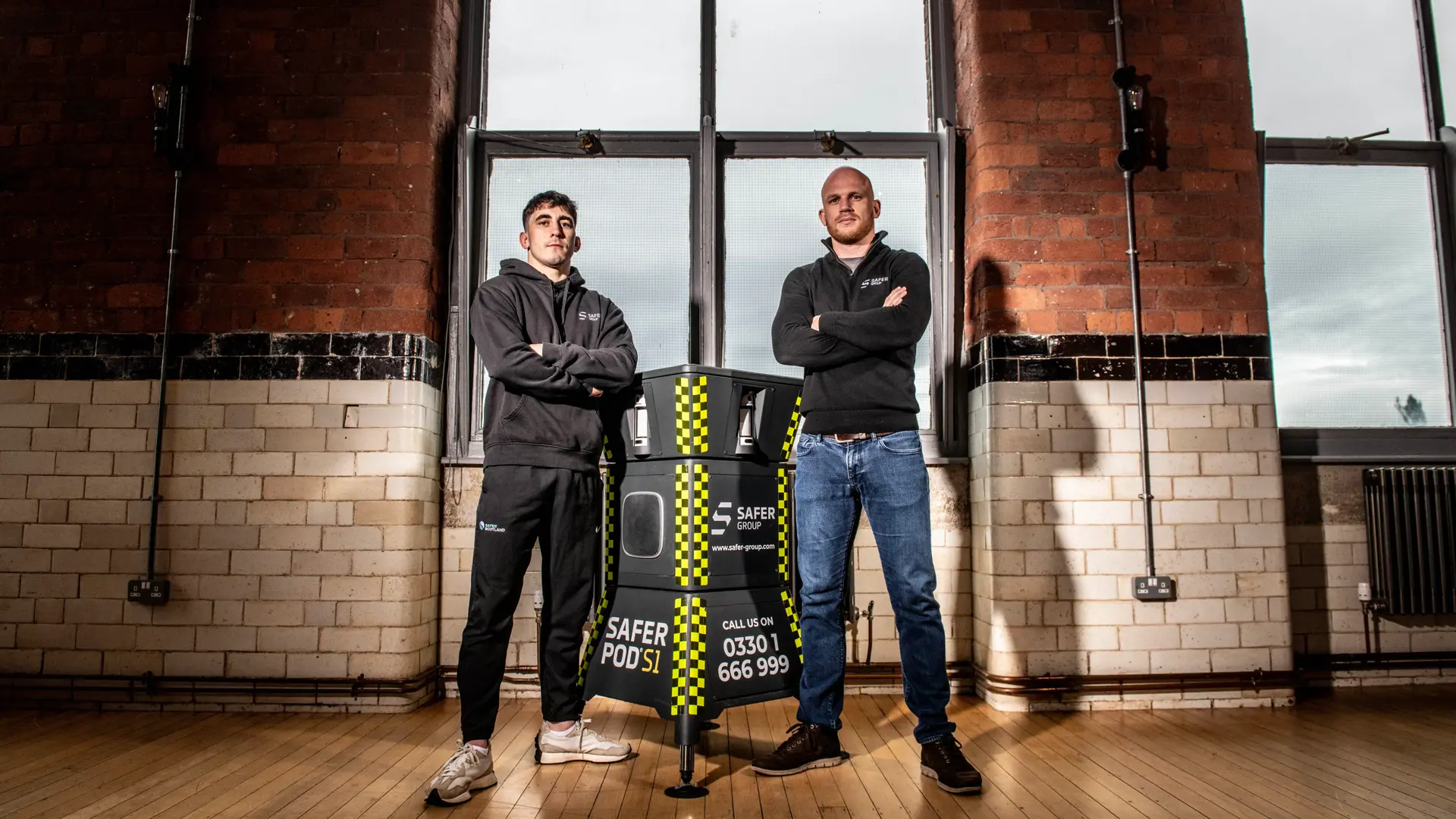 MMA fighter Reece McEwan and Group chief executive Ryan Clark stand confidently flanking a Safer Group S1 pod in a brightly lit, industrial-style interior with large windows.