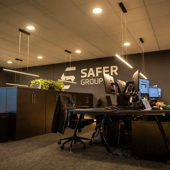 A modern office with multiple computer stations, sleek black furniture, and a large wall featuring the "SAFER GROUP" logo. An individual is seen using one of the computers.