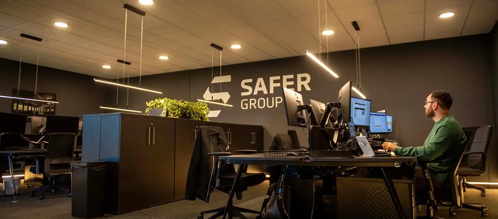 A modern office with multiple computer stations, sleek black furniture, and a large wall featuring the "SAFER GROUP" logo. An individual is seen using one of the computers.
