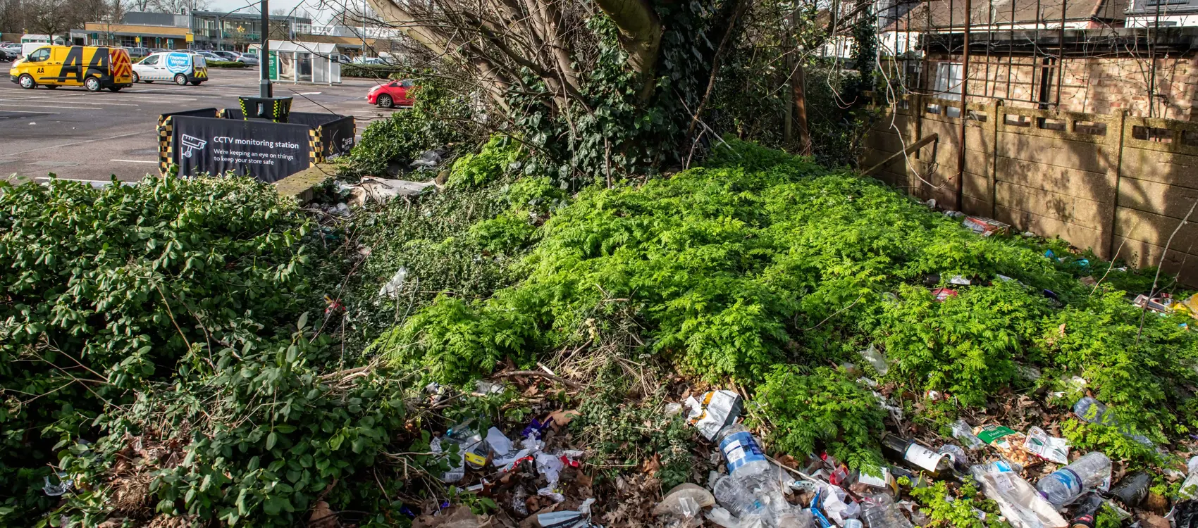 A cluttered area with overgrown greenery hiding piles of litter, including bottles and packaging, near a supermarket car park. A safer POD S7 is in the background surrounded by a safety barrier.