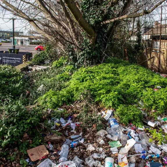 A cluttered area with overgrown greenery hiding piles of litter, including bottles and packaging, near a supermarket car park. A safer POD S7 is in the background surrounded by a safety barrier.