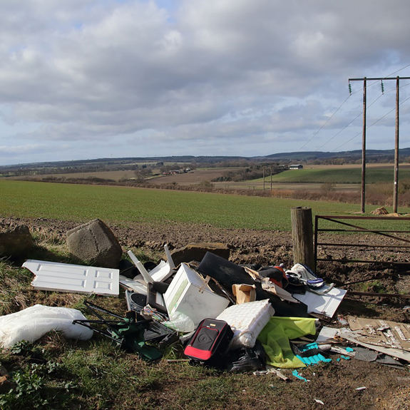 A pile of discarded household items and rubbish near an open field, with rolling hills and cloudy sky in the background.