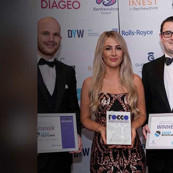 Three individuals stand together at an awards event, holding certificates and a trophy, dressed in formal attire with a backdrop of sponsor logos.