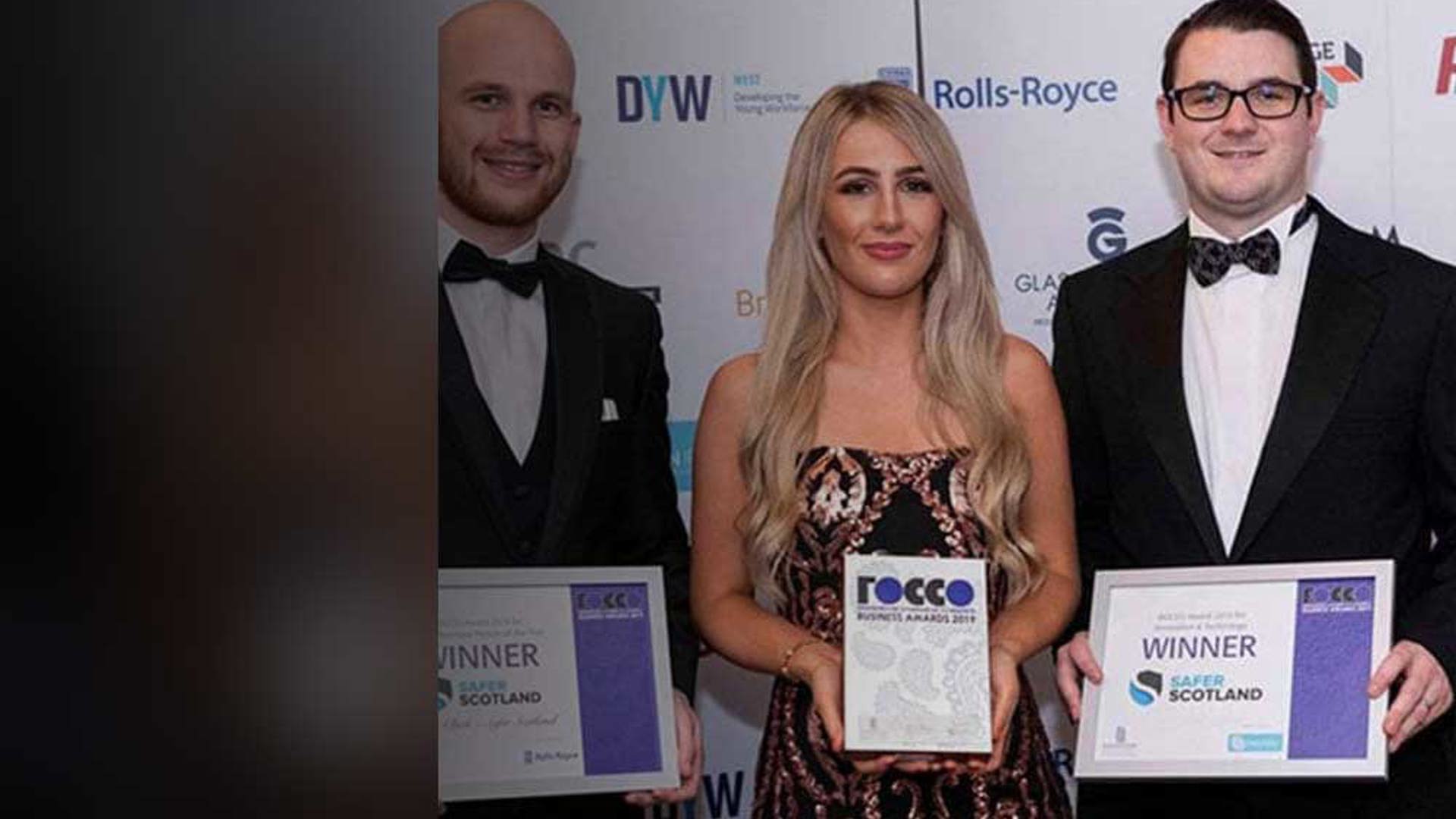 Three individuals stand together at an awards event, holding certificates and a trophy, dressed in formal attire with a backdrop of sponsor logos.