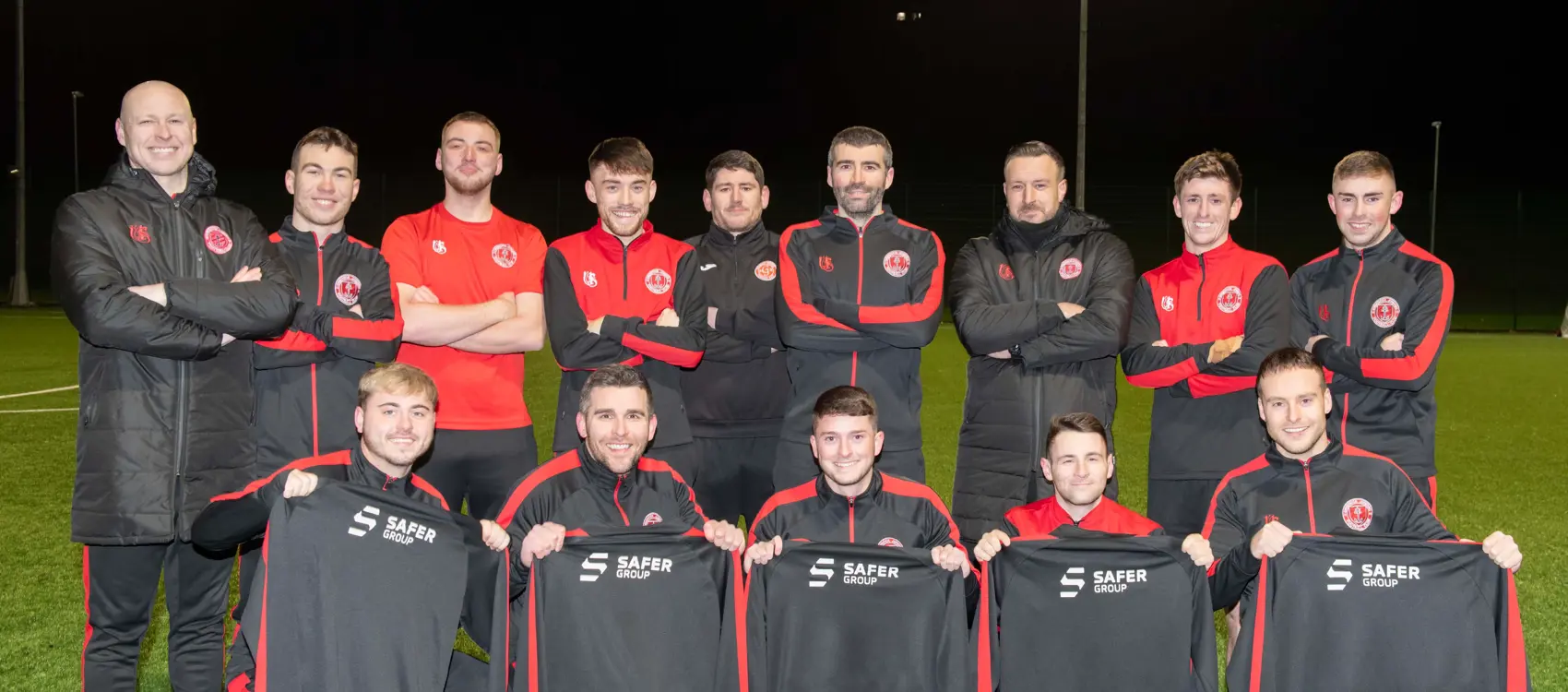 A squad of football players in dark and red team uniforms, posing on a well-lit field, holding team jackets with a safer group sponsor logo.