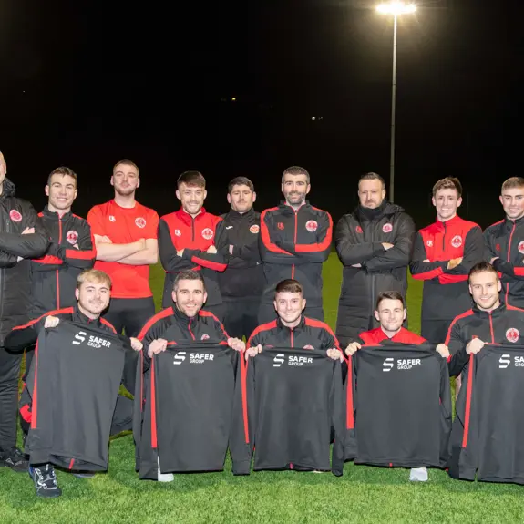 A squad of football players in dark and red team uniforms, posing on a well-lit field, holding team jackets with a safer group sponsor logo.